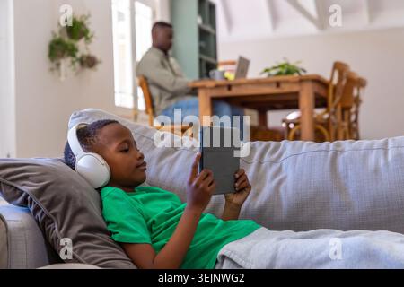 African American father and boy at home on couch using tablet with headset while working laptop. Family, remote, workspace, cozy Stock Photo