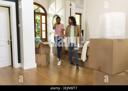Diverse couple holding hands walking through foyer surrounded by cardboard boxes and bubble wrap. Unpacking, moving, home, hallway, sunlit, hardwood, Stock Photo