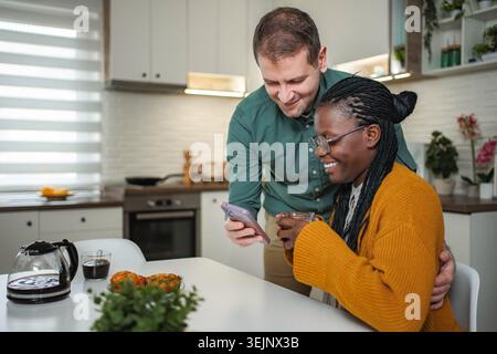 Diverse couple smiling, using smartphone, having breakfast in kitchen Stock Photo