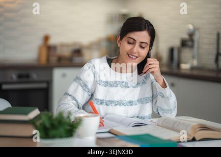 Young woman studying at home while talking on phone Stock Photo
