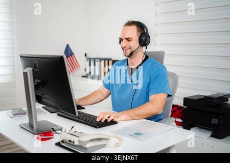 Man in scrubs smiling, using a computer and headset for remote medical work Stock Photo