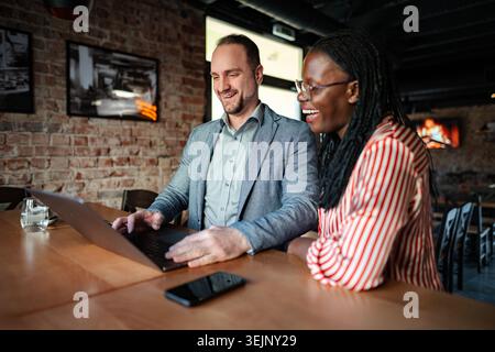 Two diverse colleagues smiling, working on a laptop together in a casual restaurant Stock Photo
