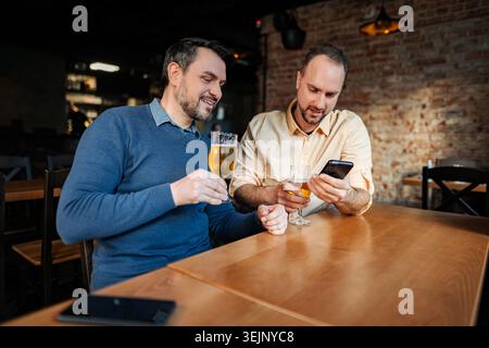 Two friends relaxing at a bar, drinking beer, using smartphone Stock Photo