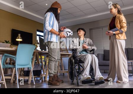 Diverse staff and client in wheelchair meeting in rehab room, man wearing beanie showing VR headset. Mobility, assistive, therapy, collaboration, demo Stock Photo
