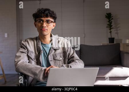 Asian man wearing light gray denim jacket and round glasses working on laptop in living room. Desk, computer, homeoffice, remote Stock Photo