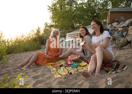 Three middle aged woman friends eat watermelon while sitting on the riverbank at a picnic Stock Photo