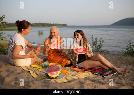 Three middle aged woman friends eat watermelon while sitting on the riverbank at a picnic Stock Photo