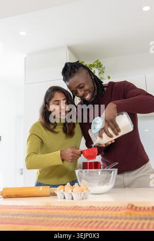 Diverse couple pouring sugar from glass jar into red measuring cup while sifting at home counter. Kitchen, baking, teamwork, col Stock Photo