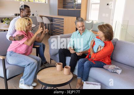 Diverse family gathering on gray sectional sofa, senior cradling swaddled infant by coffee table. Multigenerational, newborn, cozy, livingroom, openpl Stock Photo
