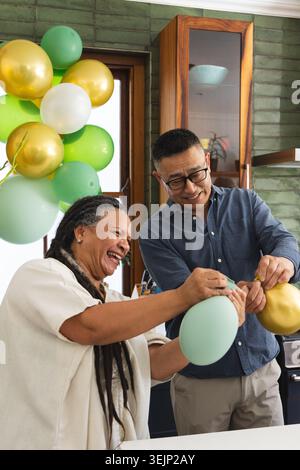 Senior Asian man, African American woman assembling green-gold-white balloon garland in kitchen. Celebration, decorating, home, Stock Photo