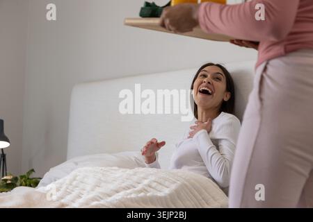 Female partners sitting in bed laughing while looking up at person holding tray with orange juice. Cozy, breakfast, morning, intimate, affection, cand Stock Photo