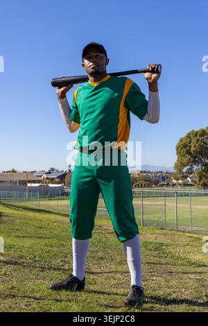 Adult African American male baseball player in green jersey black cap standing on field holding bat. Athlete, advertising, vibra Stock Photo