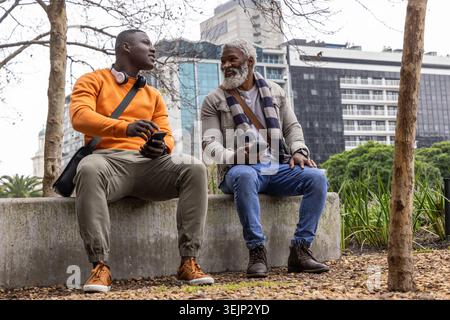 African American friends sitting and chatting on concrete bench in urban park, holding smartphones. Conversation, headphones, beard, overcast, citysca Stock Photo