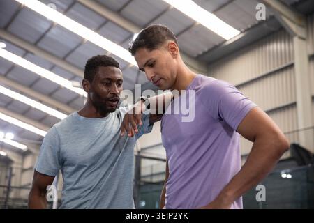 Diverse male training partners coaching and listening, standing in gym with short-sleeve shirts. Athletes, camaraderie, court, facility, industrial, n Stock Photo