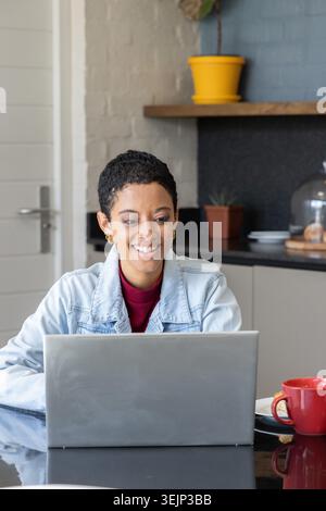 African American woman typing on silver laptop at glossy table wearing denim jacket, red mug nearby. Kitchen, homeoffice, remote Stock Photo