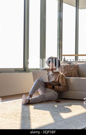 African American man sitting on floor in living room, using laptop, smartphone nearby, copy space. Homeoffice, remote, focus, pr Stock Photo