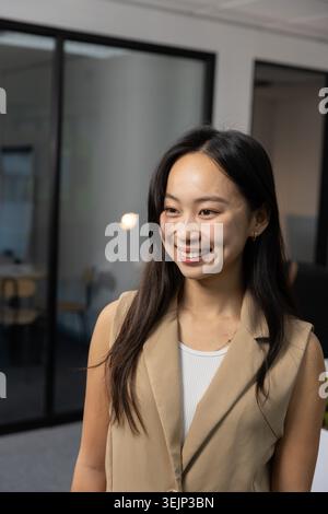 Adult Asian female standing near glass-walled room in office, smiling, wearing beige blazer. Professional, contemporary, workspa Stock Photo