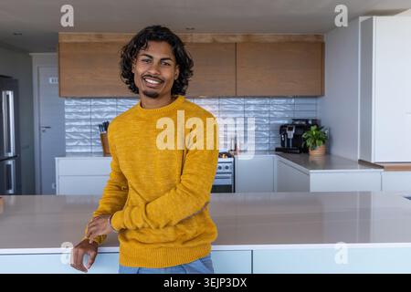 Indian man in mid 20s leaning against kitchen island in modern kitchen near coffee machine. Home, minimal, warm, contemporary, s Stock Photo