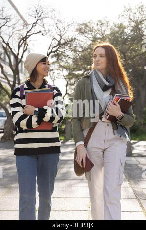 Diverse female students walking on campus path carrying notebooks, backpacks, scarf, smartphone. Outdoor, casual, friendship, co Stock Photo