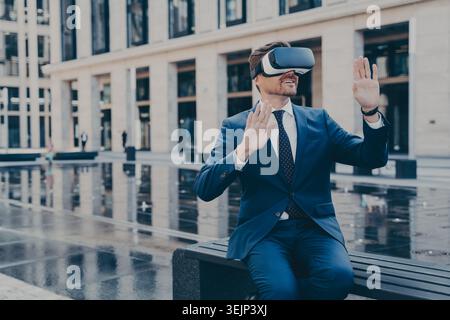 Man in VR goggles trying to reach something in virtual reality with his hands while sitting outdoors Stock Photo