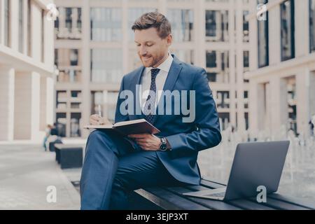 Relaxed and confident office worker in formal blue suit working outside while sitting on bench, using laptop and writing notes in agenda, city buildin Stock Photo