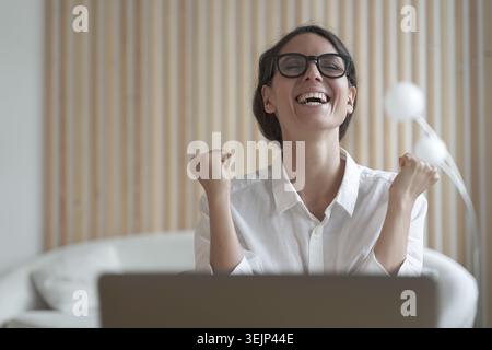Excited italian lady clenching fists with joy while sitting in front of laptop at workplace Stock Photo