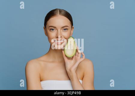 Horizontal shot of happy brunette woman holds half of avocado near face uses anti aging organic recipes stands refreshed indoor Stock Photo