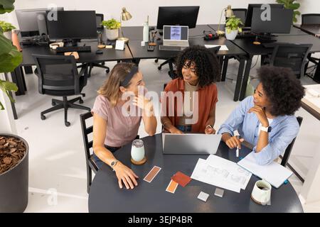 Diverse women coworkers in office wear reviewing floor plans at office table with laptop, swatches. Collaboration, design, plann Stock Photo