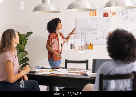 Diverse female coworkers presenting plans and reviewing swatches at modern studio table with laptop. Design, architecture, colla Stock Photo
