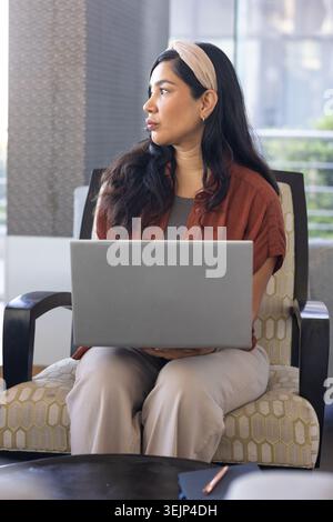 Mid-adult African American woman sitting in chair in lounge using laptop with notebook on table. Armchair, device, coworking, ur Stock Photo