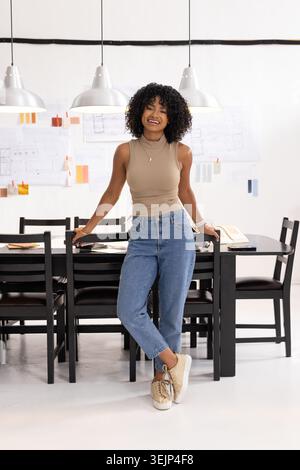 Adult female leaning against long black conference table in design studio, studying taped plans. Creative, architecture, drawing Stock Photo