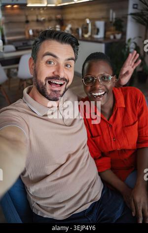 Happy multi-ethnic couple taking a selfie at home, waving at the camera. Stock Photo