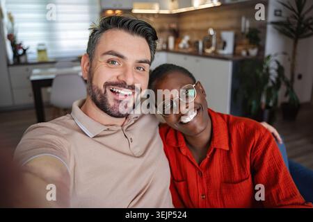 Happy multi-ethnic couple taking a selfie at home smiling at the camera Stock Photo