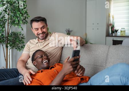 Interracial couple laughing and using smartphone on sofa at home Stock Photo
