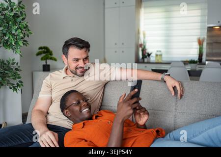 Happy multi-ethnic couple relaxing on sofa using mobile phone at home Stock Photo