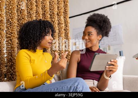 Diverse female friends discussing project on couch at studio, mustard top, burgundy top, tablet. Collaboration, brainstorming, blueprints, sketches, d Stock Photo