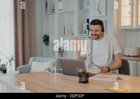 Happy male freelancer using headset and working at home during quarantine while looking at laptop screen Stock Photo