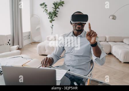 African american businessman wearing vr glasses manages business project in augmented reality at laptop. Black male businessperson working in virtual Stock Photo