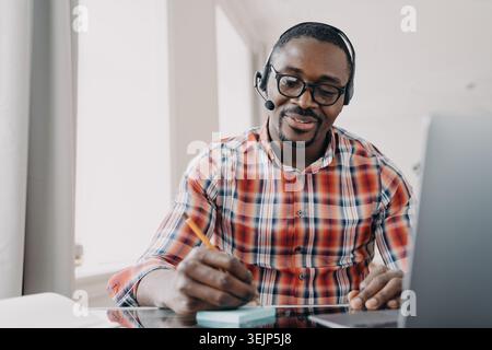 African american male student in headset makes notes, learning online at laptop. Distance education Stock Photo