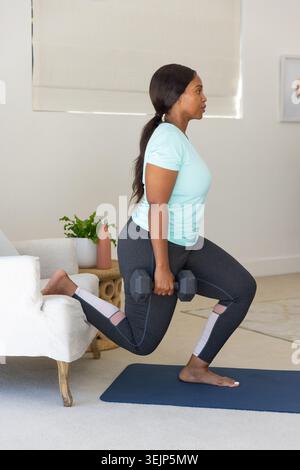 African woman doing rear-lunge on blue mat at home wearing workout top holding hex weights Stock Photo