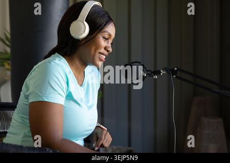 Woman wearing white headset and light blue shirt smiling while leaning toward black mic at studio Stock Photo