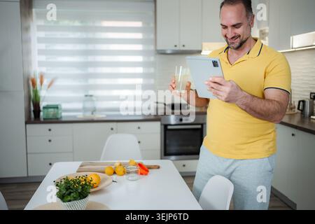 Man enjoying refreshing lemonade while using digital tablet in kitchen Stock Photo