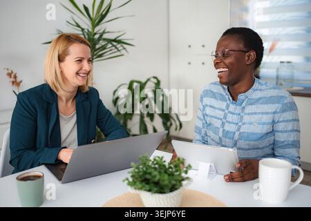 Two diverse women colleagues laughing while working with a laptop and tablet Stock Photo