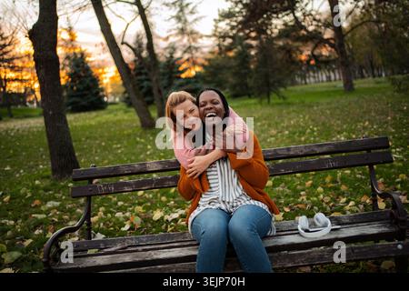 Diverse friends hugging laughing together on park bench Stock Photo