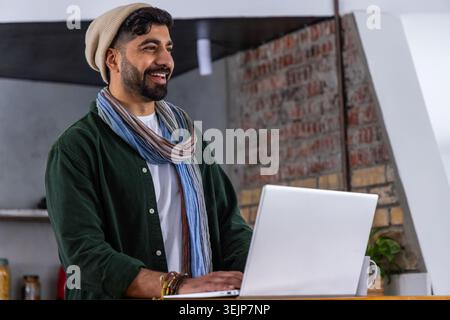 Man standing at high counter in loft workspace, wearing beanie and scarf, using laptop. Studio, brick, industrial, rustic, cozy, warm, plant Stock Photo