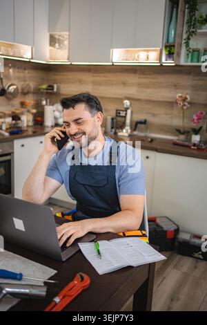 Handyman working on laptop and talking on phone in kitchen Stock Photo