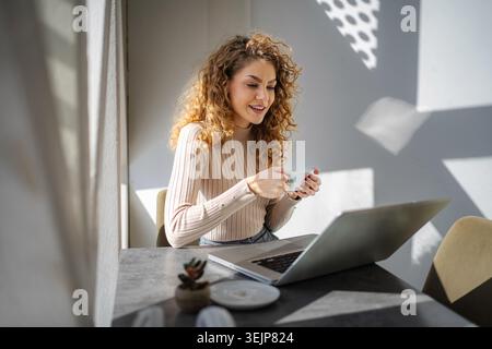Woman with curly hair using laptop while drinking coffee Stock Photo
