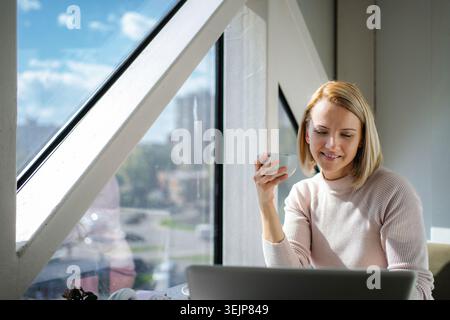 Woman drinking coffee while working on laptop by window Stock Photo