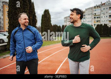 Two adult men running together on a sports track, having a conversation and smiling Stock Photo