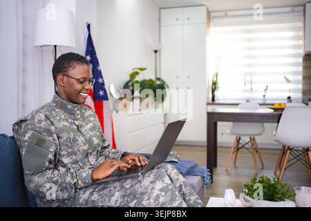 Happy female soldier in camouflage uniform is using laptop at home Stock Photo